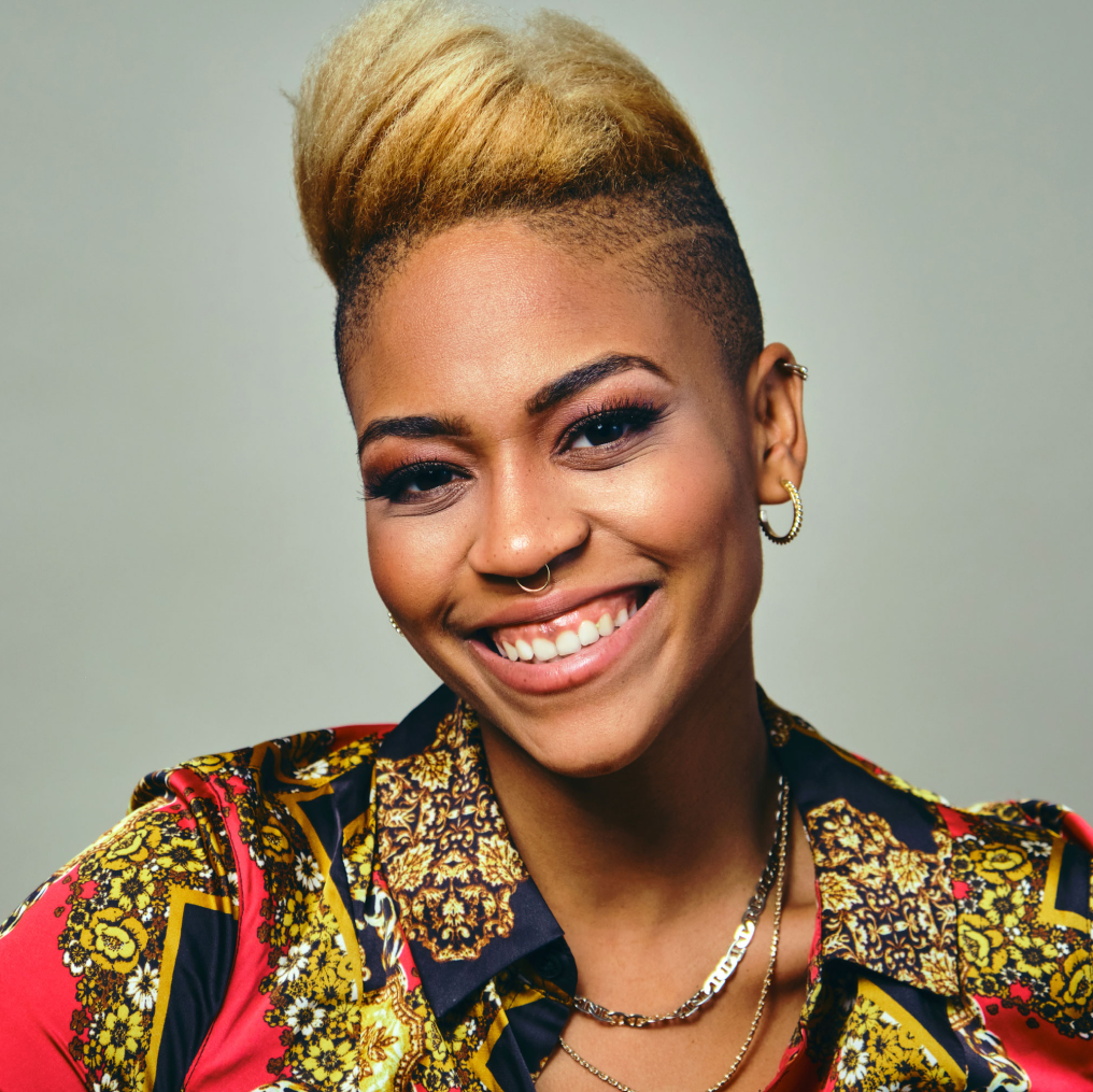 Portrait of a woman with a radiant smile, showcasing her light brown skin and a stylish undercut hairstyle with blonde highlights on top. She wears a colorful shirt adorned with bold floral and paisley patterns and is accessorized with hoop earrings, a nose ring, and a thin necklace. Her makeup includes well-defined eyebrows, eyeliner, and pink lipstick.