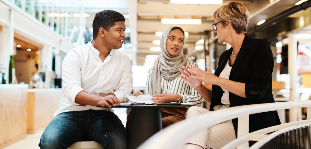 Three individuals sit at a table in a busy indoor setting. On the left, a young man with medium brown skin and short black hair, wearing a white button-up shirt and denim jeans, smiles while looking towards his companions. In the center, a young woman with olive skin and wearing a light gray hijab and white blouse, appears engaged in conversation with the woman on the right. This older woman, who has short blonde hair and wears glasses, a black blazer, and black top, gestures with her hands as she speaks. The background shows the multi-level architecture of the mall with several other visitors visible