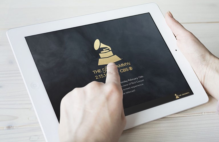 Hands holding a white tablet showing a black background with the Grammy's logo and text underneath that seems to read "The 58th Grammys", with a date and details below that are covered by the person's hand