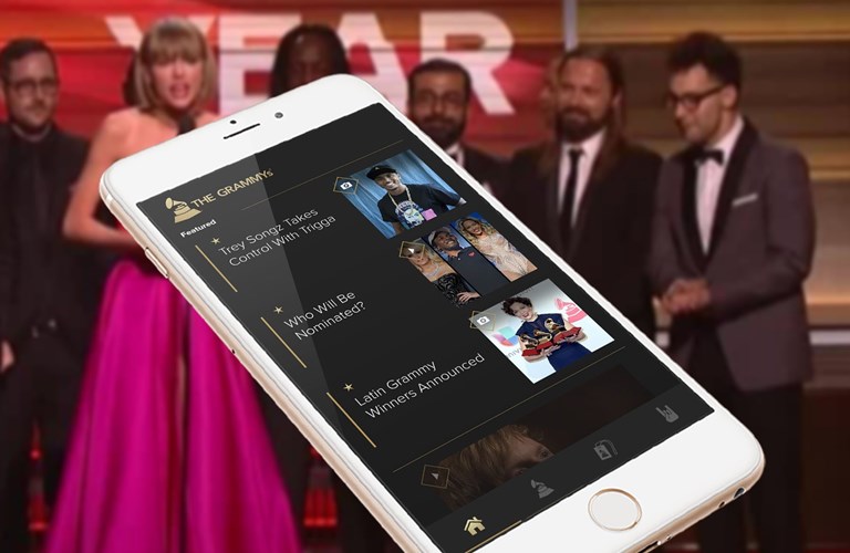 Background of Taylor Swift and others accepting a Grammy, foreground of a cellphone screen showing the home page of the app which includes a list of features news bites 