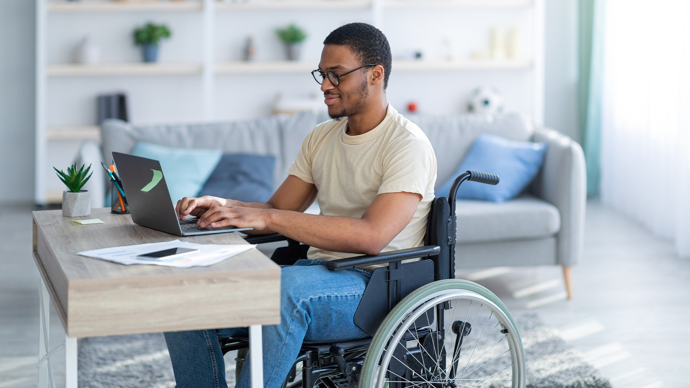 A young Black man sits in his wheelchair in a clean and well appointed apartment working on something on his laptop.