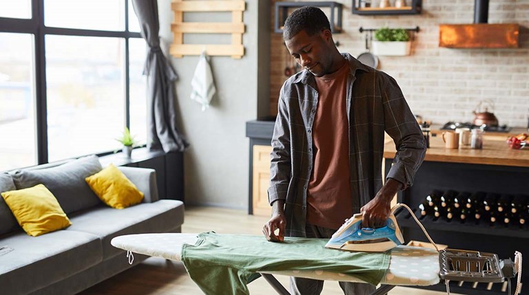 A young Black man irons a green shirt in his studio apartment