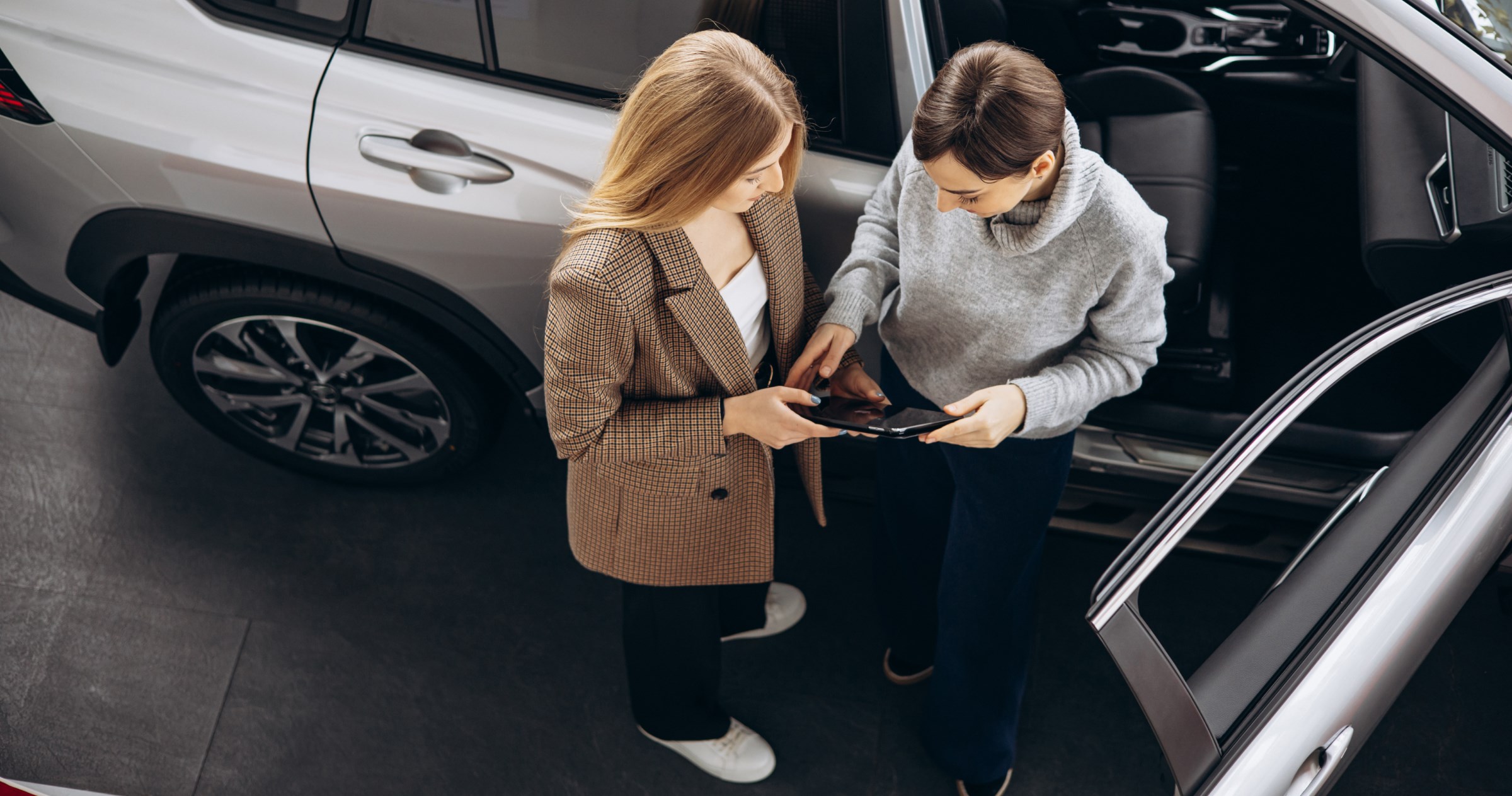 Two women stand next to an open SUV looking at a tablet.