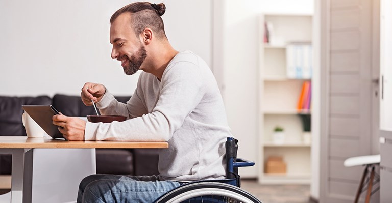 Young Caucasian man in wheelchair eating breakfast and looking at his tablet