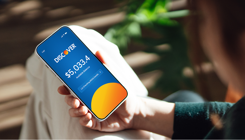 Hand holding a smartphone with the Discover banking app showing an available balance. 