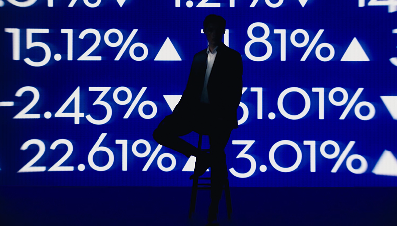 Silhouette of a person on a stool before a blue screen filled with large percentages and up/down arrows 