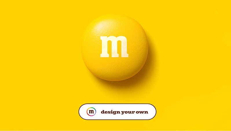 Large yellow M&M candy with white ‘m’ on a yellow background; button below reads ‘design your own. 