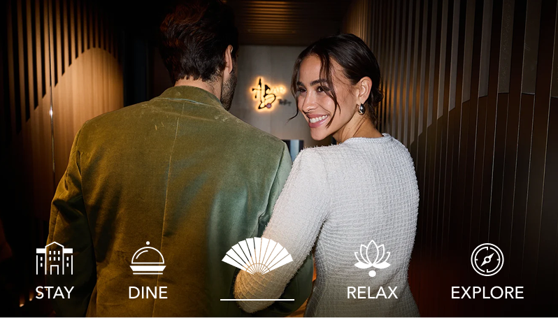 Couple walking into a dimly lit hotel corridor; woman looks back smiling. Icons: Stay, Dine, Relax, Explore, with Mandarin Oriental fan. 