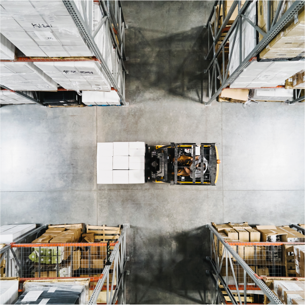 Overhead view of a forklift carrying a pallet of white boxes between tall warehouse racks. 