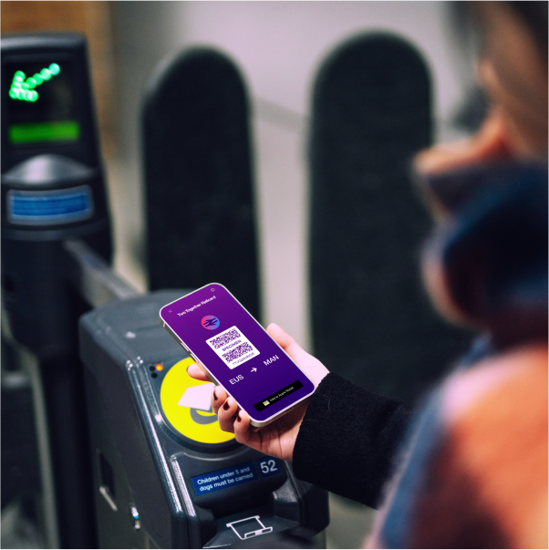 Commuter scanning a mobile QR ticket at a train station barrier—the phone shows a purple digital ticket (EUS → MAN). 