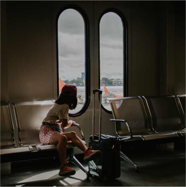 Child sitting on an airport bench with a small rolling suitcase, looking out at an airplane through tall windows. 