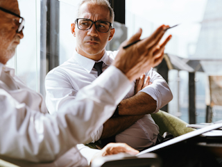 Two male executives are engaged in a focused conversation in a modern office setting. Both are wearing glasses and white dress shirts, and one man is gesturing with a pen while explaining something, while the other listens attentively with his arms crossed.