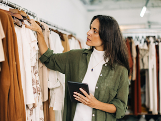 A woman in a retail store is checking the clothing inventory. She is wearing a green button-up shirt and holding a tablet as she looks carefully at the garments hanging on the rack. The store has neutral tones, and the woman is focused on her task, demonstrating technology use in retail for inventory management or digital transformation. The atmosphere is calm and organized.