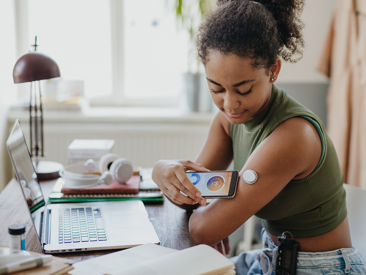 Young woman using a smartphone to scan a digital glucose monitor on her arm, showcasing modern healthtech solutions for diabetes management in a home setting.
