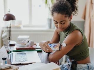 Young woman using a smartphone to scan a digital glucose monitor on her arm, showcasing modern healthtech solutions for diabetes management in a home setting.