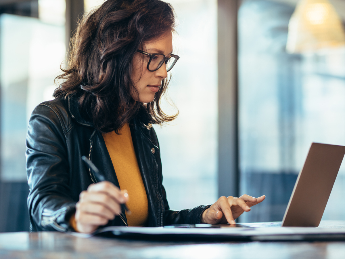 A woman wearing glasses and a leather jacket working on a laptop, focused on the screen, with a pen in her hand, in a professional setting.
