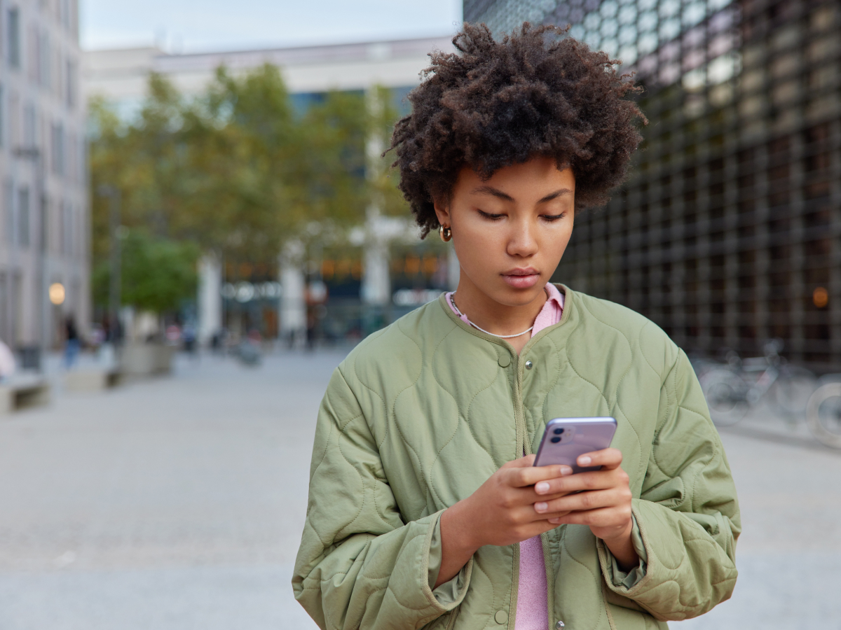 A young woman with curly hair, wearing a green jacket, looks intently at her smartphone while standing in a modern city environment.