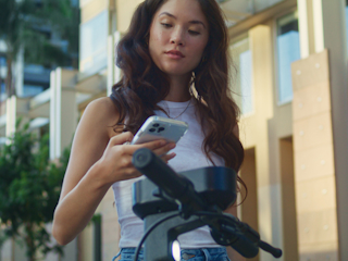 A young woman with long wavy hair is standing outdoors in a city environment, looking down at her smartphone.