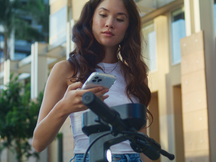 A young woman with long wavy hair is standing outdoors in a city environment, looking down at her smartphone.