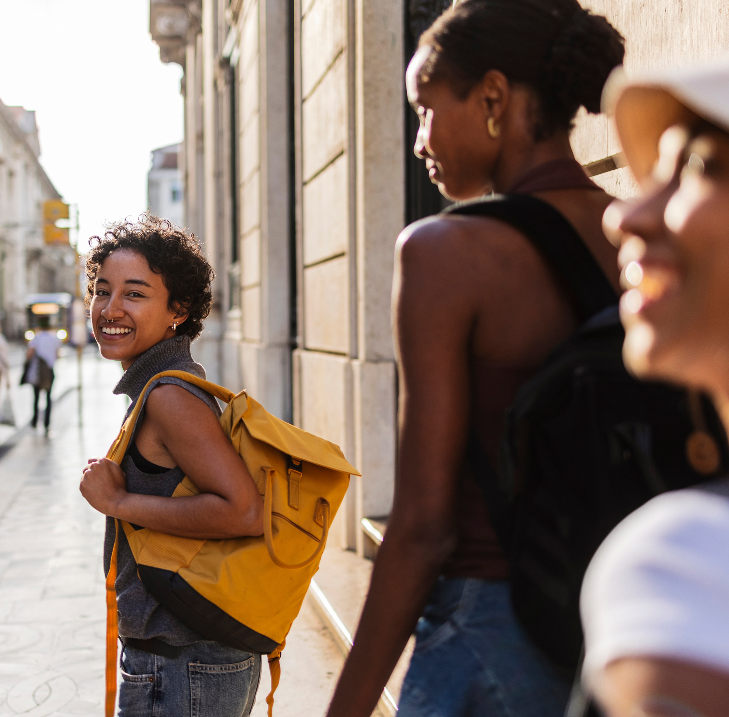 three women walking down a sunlit city street. The woman in the foreground is smiling warmly at the camera and carrying a bright yellow backpack, creating a vibrant and inviting travel vibe.