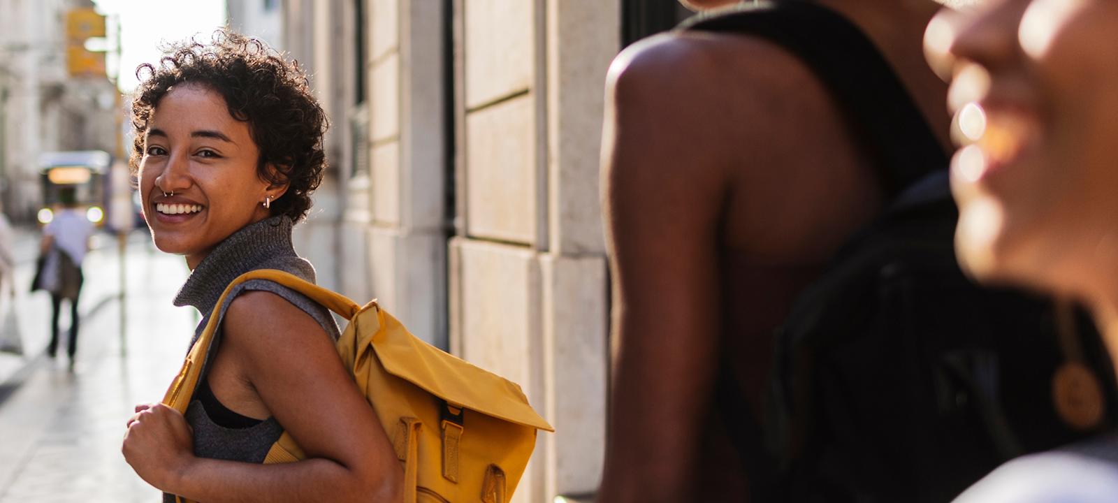three women walking down a sunlit city street. The woman in the foreground is smiling warmly at the camera and carrying a bright yellow backpack, creating a vibrant and inviting travel vibe.