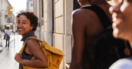 three women walking down a sunlit city street. The woman in the foreground is smiling warmly at the camera and carrying a bright yellow backpack, creating a vibrant and inviting travel vibe.
