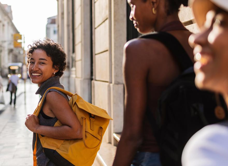 three women walking down a sunlit city street. The woman in the foreground is smiling warmly at the camera and carrying a bright yellow backpack, creating a vibrant and inviting travel vibe.