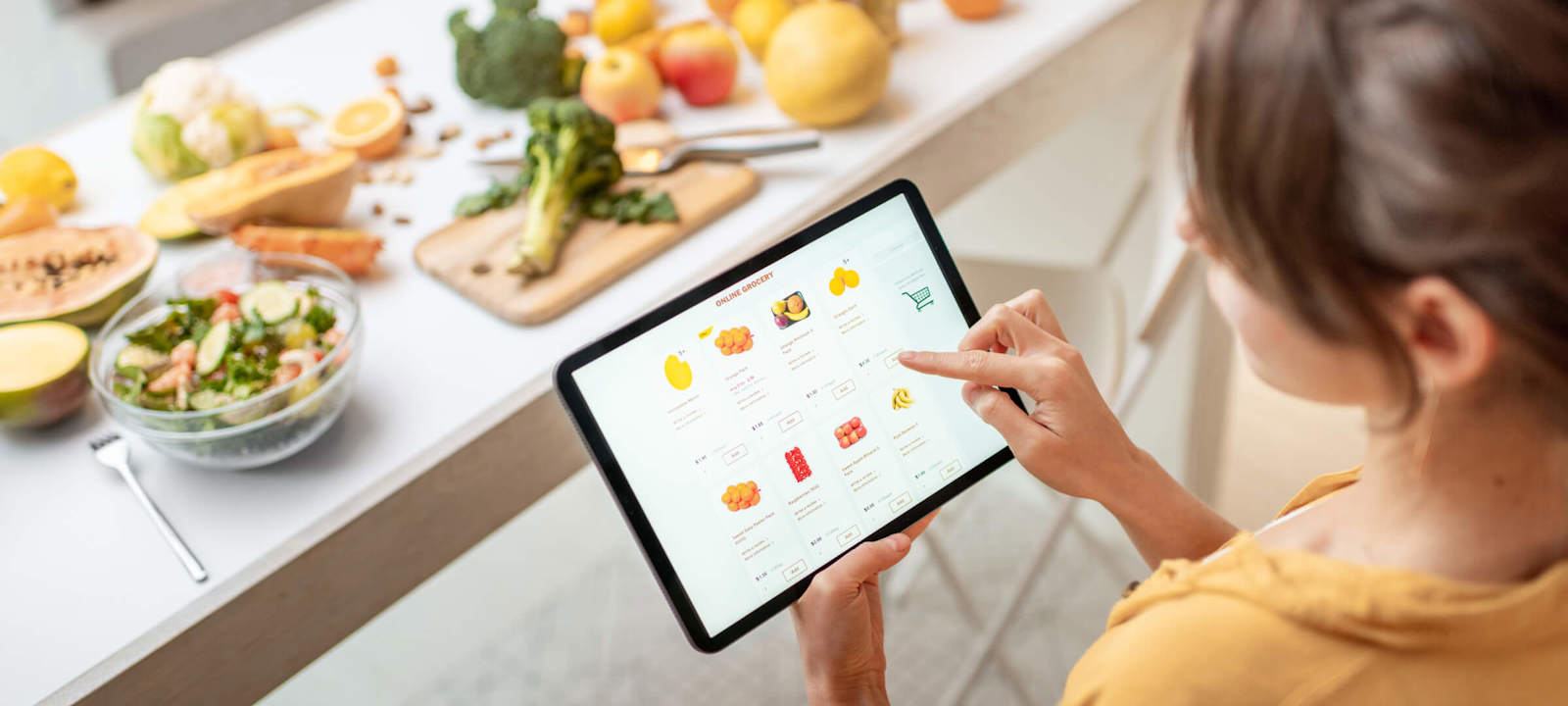A woman using a tablet to shop for groceries online while standing in a kitchen. The kitchen counter is filled with fresh fruits, vegetables, and a salad, creating a healthy and vibrant atmosphere. The tablet screen displays various grocery items, emphasizing the convenience of online grocery shopping.