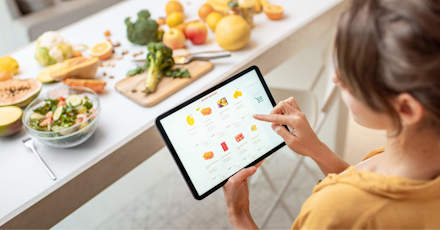 A woman using a tablet to shop for groceries online while standing in a kitchen. The kitchen counter is filled with fresh fruits, vegetables, and a salad, creating a healthy and vibrant atmosphere. The tablet screen displays various grocery items, emphasizing the convenience of online grocery shopping.