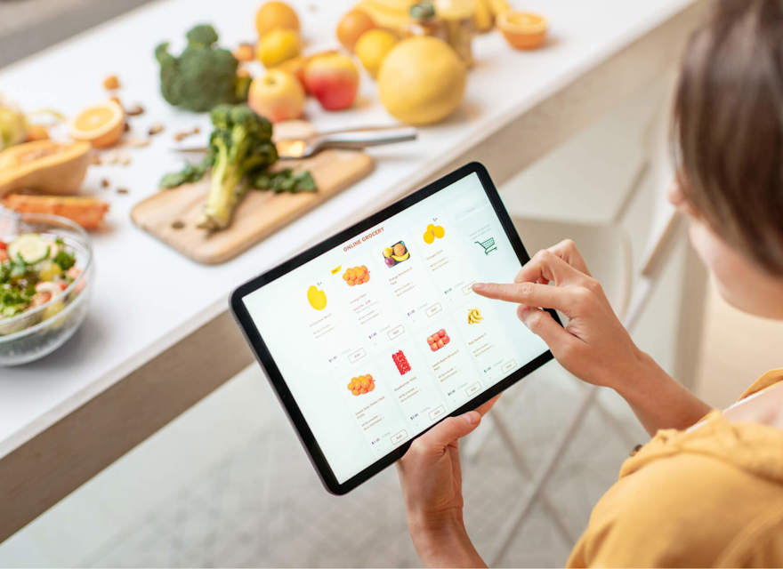 A woman using a tablet to shop for groceries online while standing in a kitchen. The kitchen counter is filled with fresh fruits, vegetables, and a salad, creating a healthy and vibrant atmosphere. The tablet screen displays various grocery items, emphasizing the convenience of online grocery shopping.