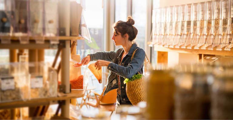 A woman shopping in a bulk food store, filling her reusable bag with dried goods from dispensers. She is carrying a woven basket with fresh produce and is focused on selecting her items. The scene is bathed in warm sunlight, emphasizing an eco-friendly, sustainable shopping experience.
