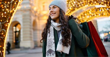 A smiling woman dressed in winter clothing, holding shopping bags while walking under festive holiday lights. She is wearing a knit hat, scarf, and coat, enjoying a seasonal shopping experience in a brightly lit, decorated urban setting.