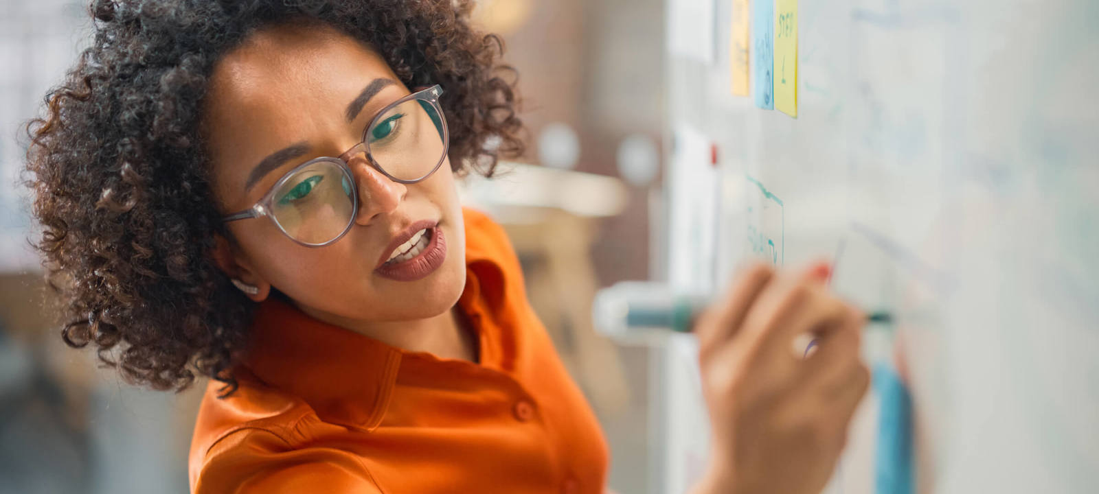 A focused woman with curly hair and glasses is writing on a whiteboard during a brainstorming or planning session. Colorful sticky notes and diagrams are visible on the board, suggesting an organized and collaborative work process. The image captures the energy and creativity of a modern professional environment, highlighting problem-solving and strategic thinking.