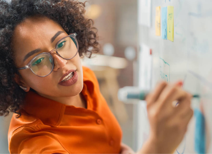 A focused woman with curly hair and glasses is writing on a whiteboard during a brainstorming or planning session. Colorful sticky notes and diagrams are visible on the board, suggesting an organized and collaborative work process. The image captures the energy and creativity of a modern professional environment, highlighting problem-solving and strategic thinking.