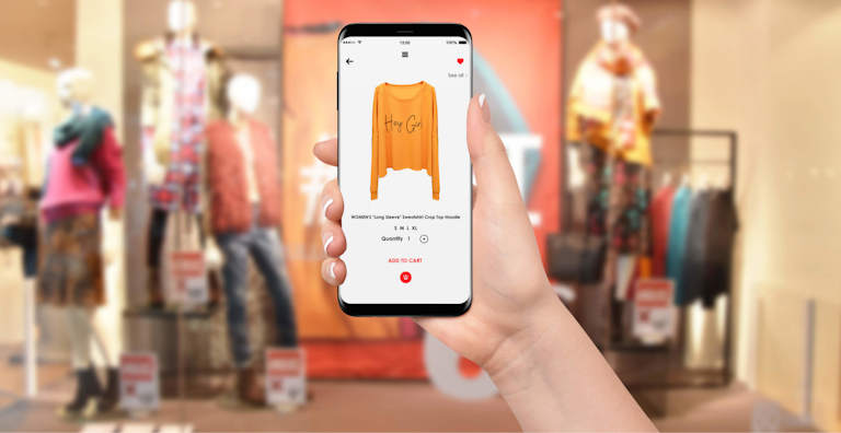A hand holding a smartphone shows an online shopping app displaying a women’s “Hey Girl” sweatshirt for purchase. In the background, a blurred retail store window showcases mannequins dressed in colorful outfits, reinforcing the connection between physical and digital shopping experiences. The interface on the phone highlights product details such as size options, quantity, and the “Add to Cart” button.