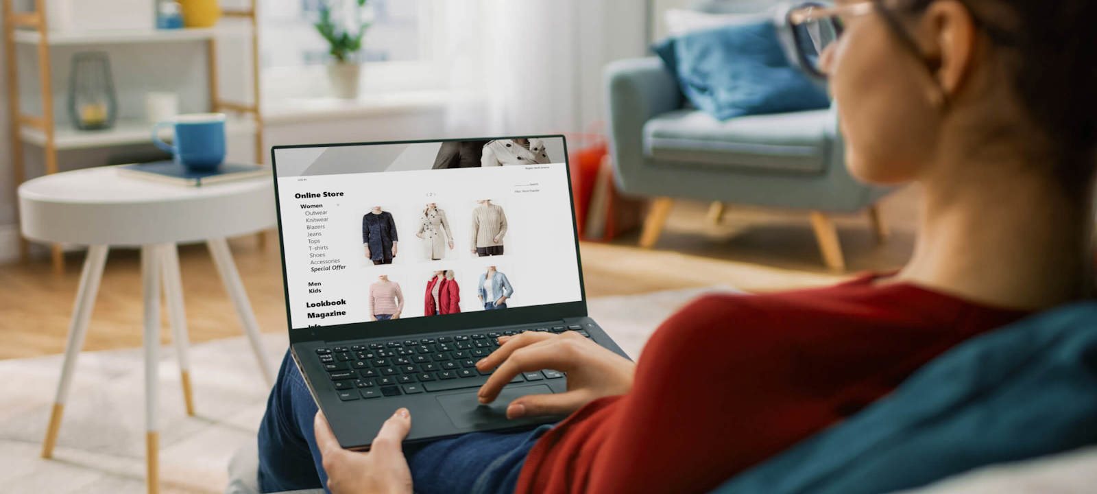 A woman sits comfortably on a sofa, browsing an online clothing store on her laptop. The screen displays various categories such as women’s clothing, accessories, and offers, along with images of different clothing items like jackets and sweaters. The cozy living room setting includes a coffee table, mug, and background furnishings, creating a relaxed shopping environment.