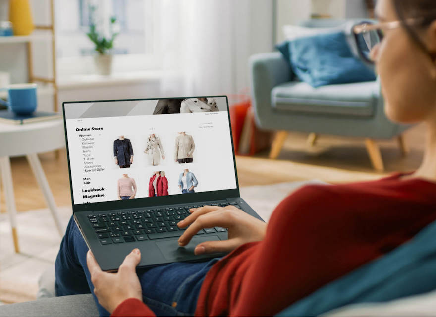 A woman sits comfortably on a sofa, browsing an online clothing store on her laptop. The screen displays various categories such as women’s clothing, accessories, and offers, along with images of different clothing items like jackets and sweaters. The cozy living room setting includes a coffee table, mug, and background furnishings, creating a relaxed shopping environment.