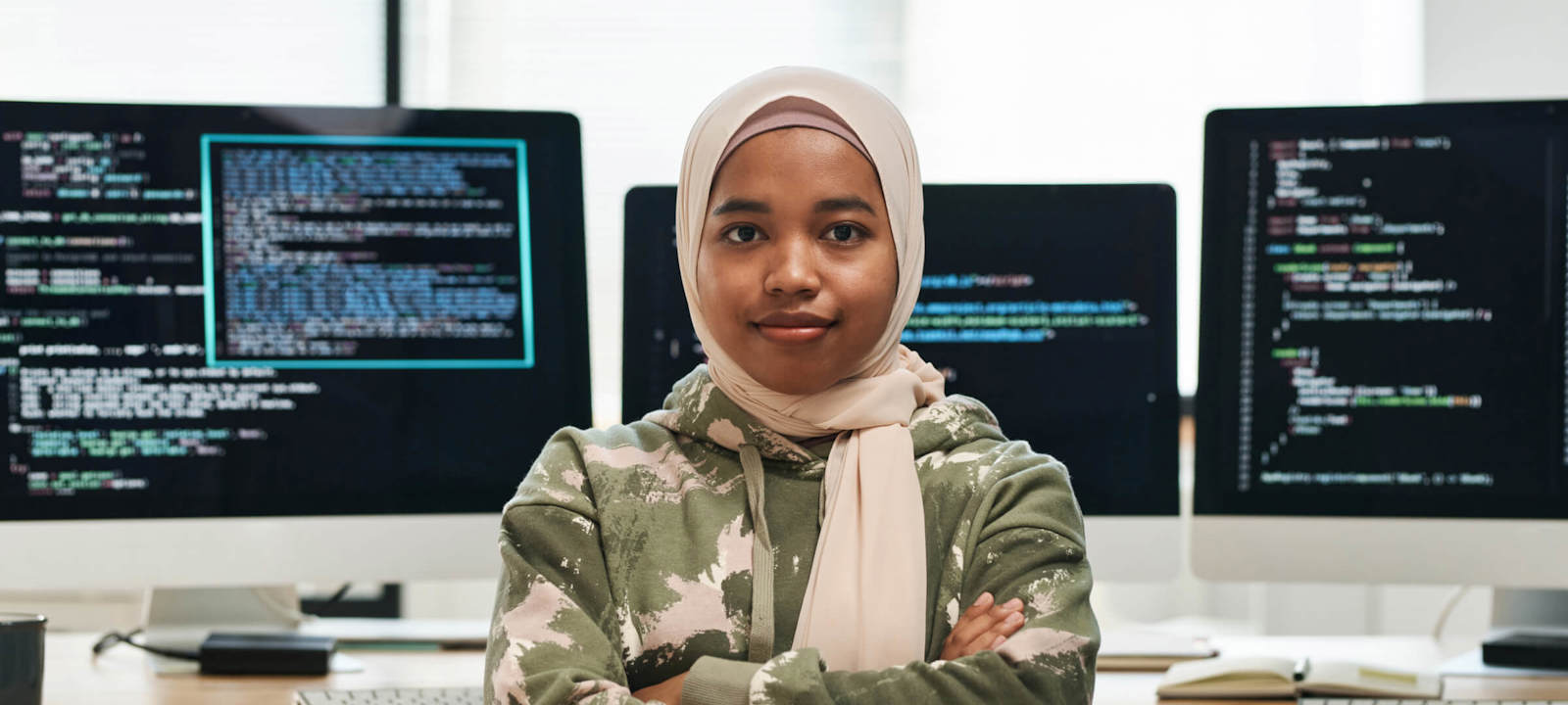 Confident software developer wearing a hijab, standing in front of computer screens with coding displayed
