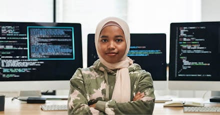 Confident software developer wearing a hijab, standing in front of computer screens with coding displayed