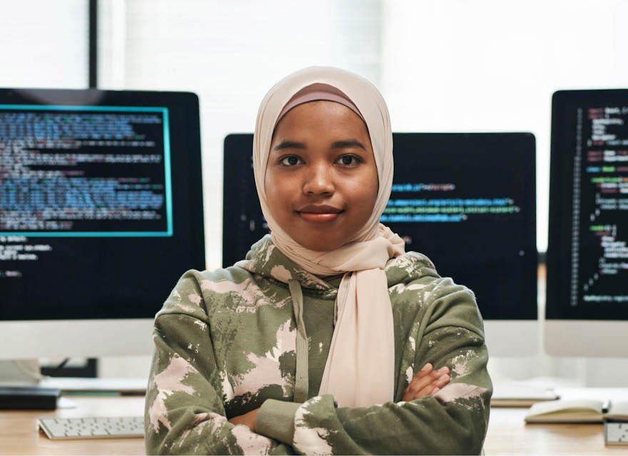 Confident software developer wearing a hijab, standing in front of computer screens with coding displayed