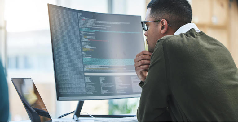 A man sitting at a desk, closely analyzing code on a large monitor. He is wearing glasses and appears focused, with a tablet placed next to the monitor. The scene reflects a high-tech work environment, possibly related to programming or cybersecurity.