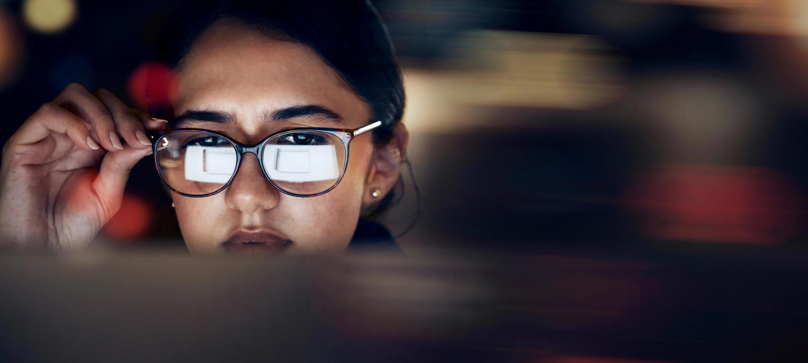 A close-up of a woman wearing glasses, intensely focused on a screen, with the reflection of the screen visible in her lenses. The background is blurred, highlighting her concentration and the digital nature of her work.