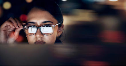 A close-up of a woman wearing glasses, intensely focused on a screen, with the reflection of the screen visible in her lenses. The background is blurred, highlighting her concentration and the digital nature of her work.