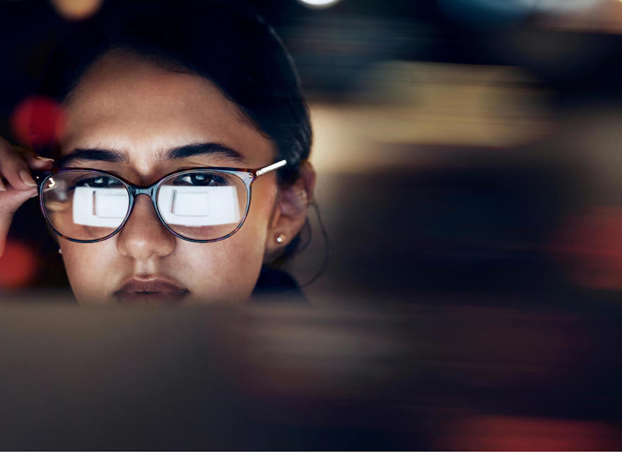 A close-up of a woman wearing glasses, intensely focused on a screen, with the reflection of the screen visible in her lenses. The background is blurred, highlighting her concentration and the digital nature of her work.