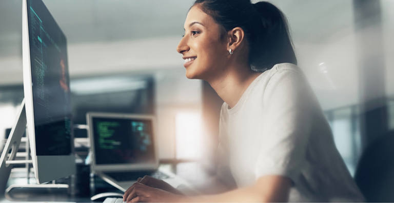 A woman smiling while working at a computer in a modern office. She is focused on the screen, which displays lines of code, with a second monitor visible in the background. The image conveys a positive work environment and engagement in programming or cybersecurity tasks.