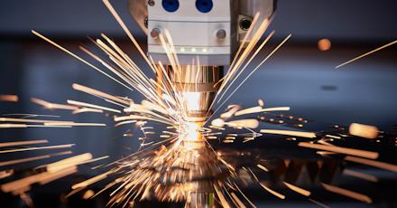 A close-up view of a laser cutting machine in action, emitting bright, golden sparks as it cuts through metal with precision. The machinery is detailed, with sharp focus on the nozzle and spark trails, creating a dynamic industrial scene.