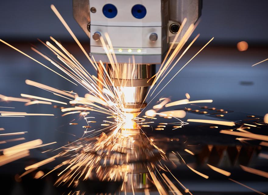 A close-up view of a laser cutting machine in action, emitting bright, golden sparks as it cuts through metal with precision. The machinery is detailed, with sharp focus on the nozzle and spark trails, creating a dynamic industrial scene.