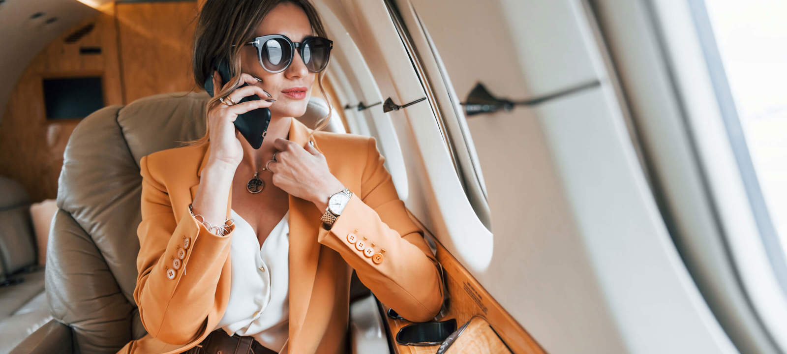 A stylish businesswoman wearing a light orange suit and large sunglasses is sitting in a private jet, talking on her phone. She looks composed and confident, with the luxurious interior of the jet visible around her. The image captures a moment of professional engagement during travel, highlighting themes of success, luxury, and mobility in a business context.
