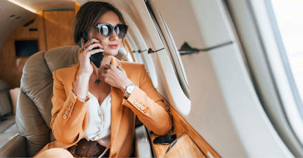 A stylish businesswoman wearing a light orange suit and large sunglasses is sitting in a private jet, talking on her phone. She looks composed and confident, with the luxurious interior of the jet visible around her. The image captures a moment of professional engagement during travel, highlighting themes of success, luxury, and mobility in a business context.