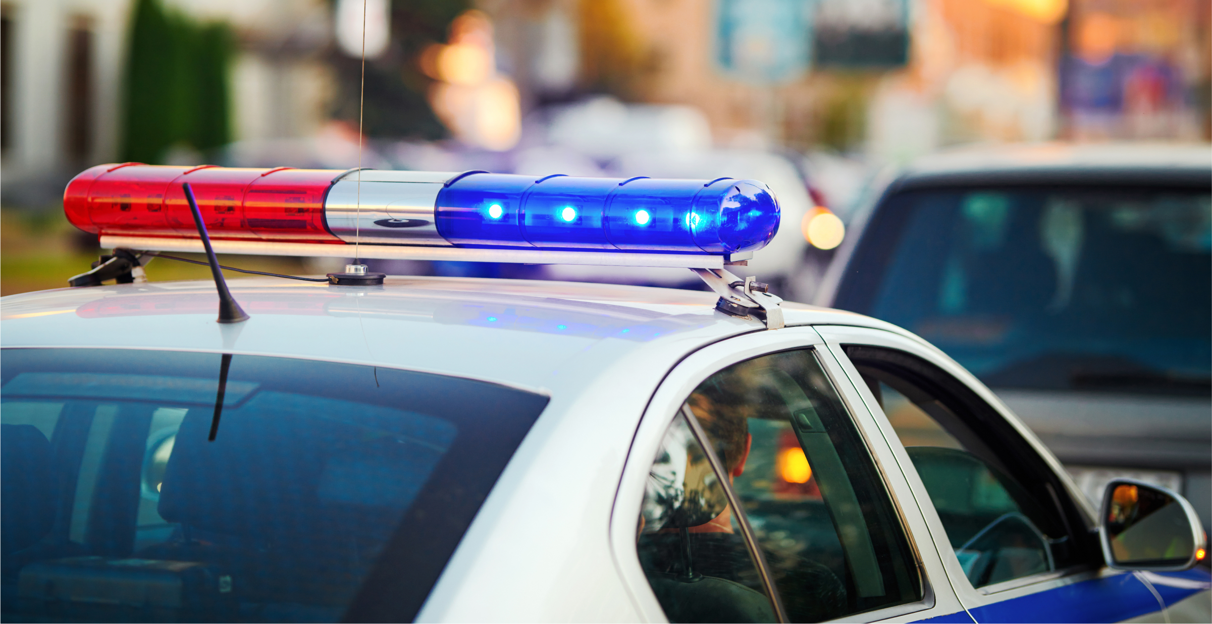 A close-up of a police car’s roof with its red and blue lights flashing. The background shows a blurred city street with other vehicles, suggesting the police car is responding to an incident or patrolling in an urban area. The image captures the sense of urgency and authority associated with law enforcement in action.